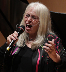 Cathy Jensen Hole singing. She is holding a mic and has long blond hair, bangs and glasses