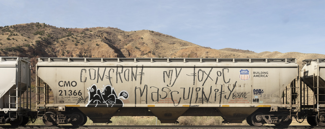 Image of a train car in front of a barren, hilly backdrop. There is graffiti on the train car, the most prominent display is the phrase "Confront my toxic masculinity."
