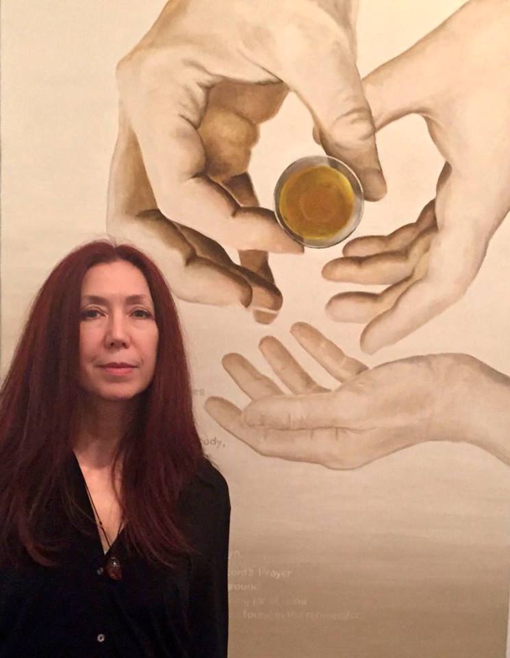 Photo of curator Linda Griggs posed in front of a painting. She is wearing a black sweater, has dark red hair past her shoulders, and is looking directly at the camera. The painting behind her is of three hands, two larger adult hands holding what looks like a small glass of brown liquid, and one smaller child-like hand. The child-like hand is open, as if ready to receive the glass from the adult's hands.
