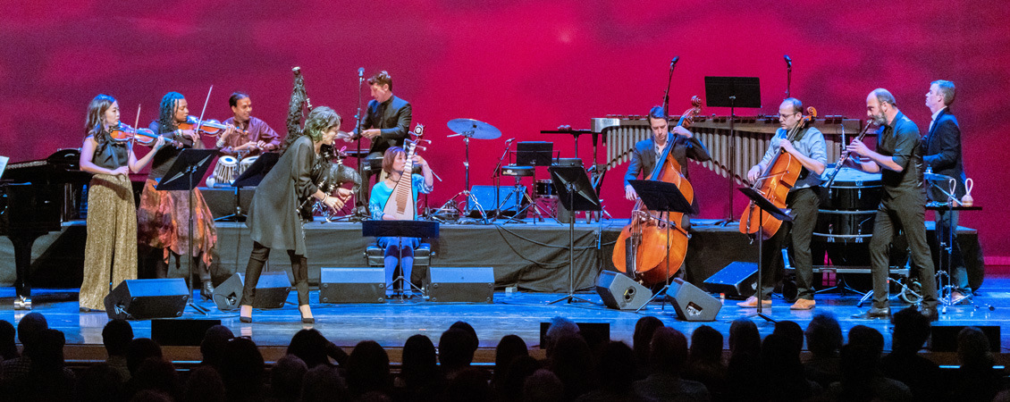Image of the Silk Road Ensemble performing on a stage. Pictured are ten musicians playing a variety of instruments, including violin, saxophone, drums, and bass. The audience members' heads are visible across the bottom of the image.