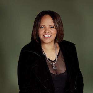 Image of a smiling individual with brown, bobbed hair, wearing a long necklace and dressed in black clothing 