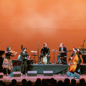 Image of the Silkroad Ensemble playing on a stage. Pictured are five musicians, left to right: a violist, a big pipe player, two drummers, and a bassist. They are playing on a stage in front of a bright orange backdrop. 
