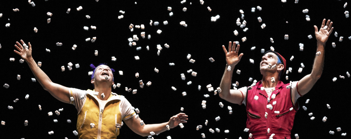 Photograph of a person against a black background, with packaging peanuts scattered in the air around them. The person is looking upwards and smiling, with arms extended above him.