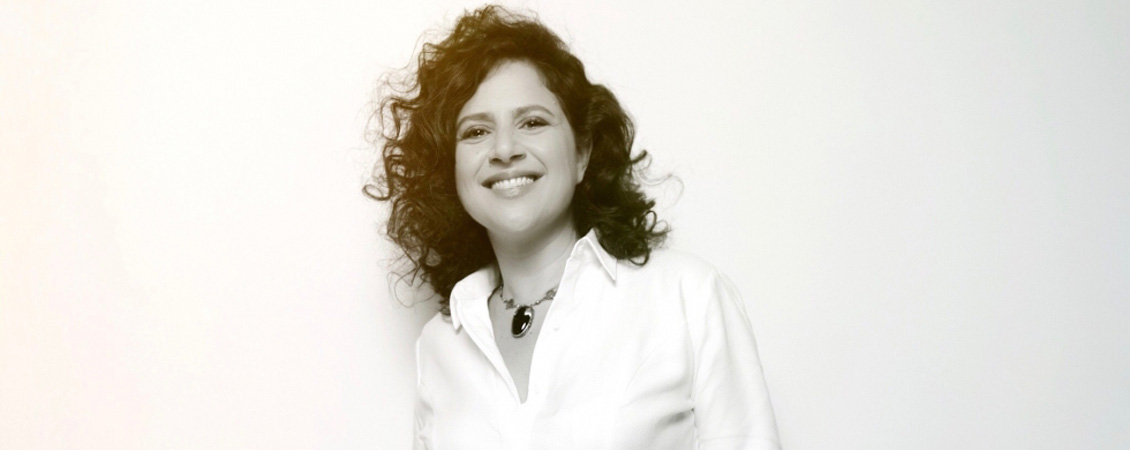 Black and white photo of Anat Cohen posing against a white background. She is wearing a white collared shirt, large oval pendant necklace. She is smiling at the camera.