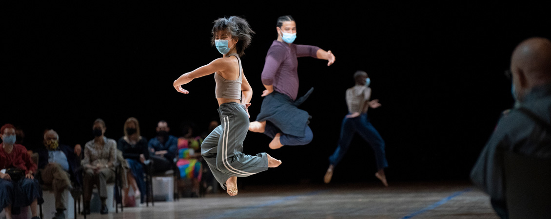 Photograph of two dancers wearing comfortable clothing and blue masks in the middle of a jump. Audience members are visible in the background sitting in chairs and socially distancing. Another dancer is also visible further in the background, mid jump. The background is pitch black.