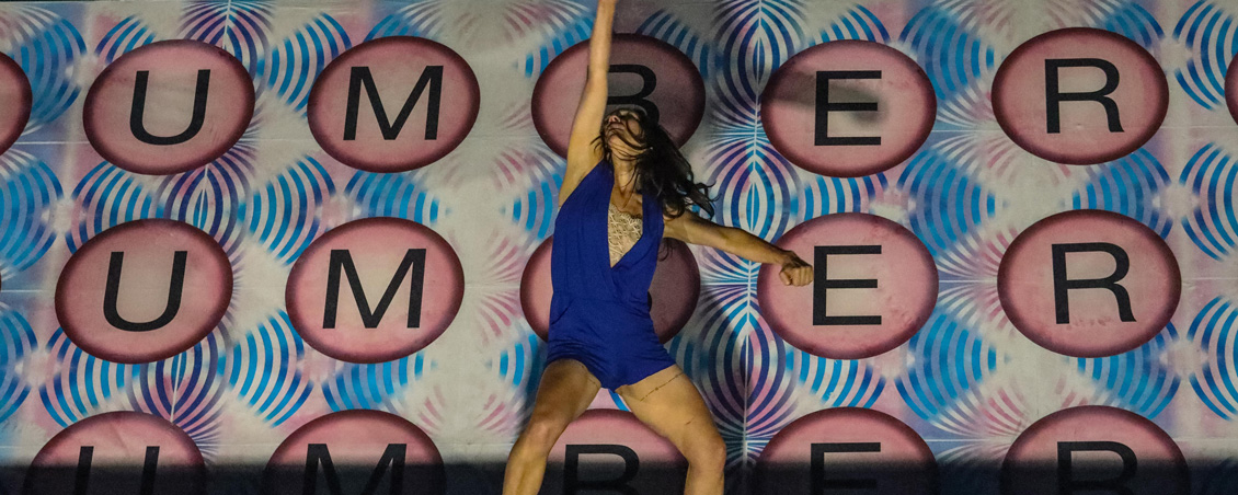 Photo of Emily Johnson on stage, with a colorful pink, white, and blue background. She is wearing a dark blue romper. She is in the middle of a performance; her head is thrown back, and her arms are stretched out, one above her head and one to her side.
