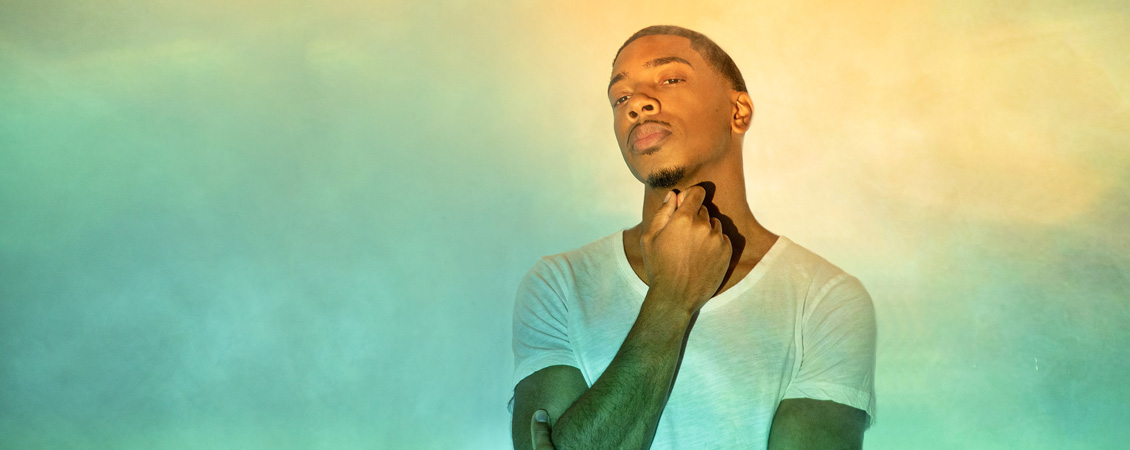 An image of Christian Sands against a yellow-green backdrop. His right hand is hovering under his chin, and he is looking at the camera with a serious, relaxed expression. 