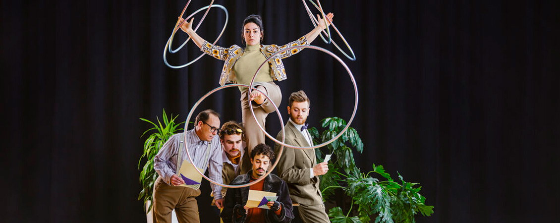 A man is sitting in a chair, holding a piece of paper and reading it. Three other men are posed around him, also peering at the paper. A woman is perched behind them, one leg raised and swinging six hoops around both arms and her leg. A few green potted plants are behind them and they are on a stage with a black curtain.