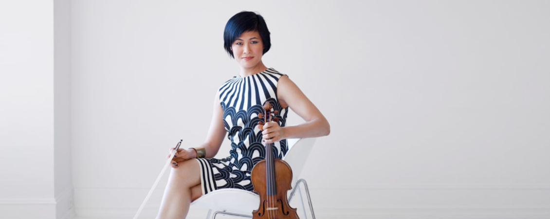 Photograph of Jennifer Koh sitting in a white chair against a white backdrop, looking directly at the camera. She has short black hair and is wearing a black and white sleeveless dress. She is holding her violin in one hand.