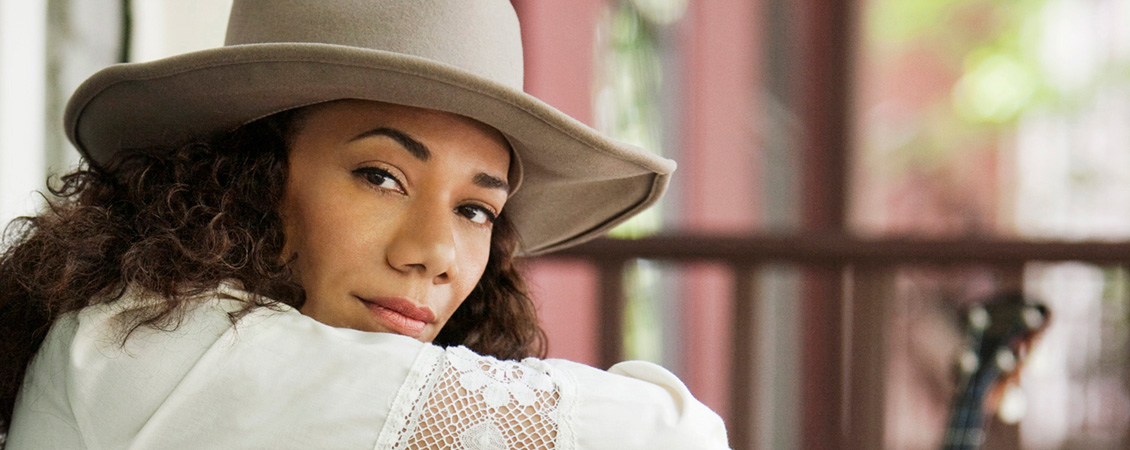 Photo of Martha Redbone. She is resting her chin on her arm and looking directly at the camera. She is wearing a lacy, white sundress and a beige cowboy hat.