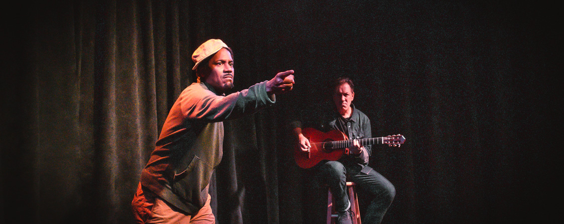 A grainy photo of two men on stage against a black curtain. One is sitting on a tall stool and playing a guitar. The other is standing to his left and pointing into the distance. He has a serious expression on his face.