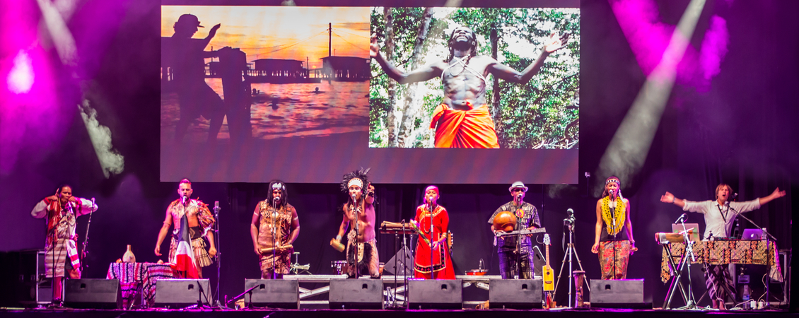 Six performers are on stage in front of a big audience. They appear to be wearing various traditional clothing and headdresses. A projection of images is shown behind them. A glowing purple light illuminates the audience and the stage,
