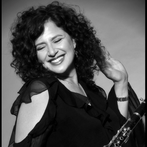 A black and white portrait photograph of Anat Cohen. She has curly, shoulder-length dark hair and is wearing a sleeveless black top. She is holding her clarinet slightly out of frame, and she is smiling widely with her eyes closed.