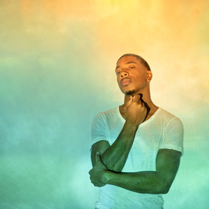 An image of Christian Sands against a yellow-green backdrop. His right hand is hovering under his chin, and he is looking at the camera with a serious, relaxed expression. 