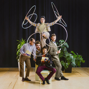 A man is sitting in a chair, holding a piece of paper and reading it. Three other men are posed around him, also peering at the paper. A woman is perched behind them, one leg raised and swinging six hoops around both arms and her leg. A few green potted plants are behind them and they are on a stage with a black curtain.