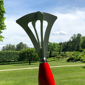 One of Peter Dellert's sculptures outdoors on a sunny, slightly cloudy day. A vintage pastry blender with a red handle is handle-side down on the grass. 