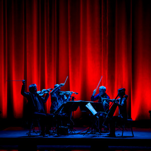 A string quartet on a small platform stage, against a vibrant red curtain backdrop. The quartet is in shadow with a faint blue light illuminating them.
