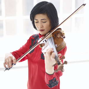 Photograph of Jennifer Koh playing the violin. Her hair is short and black, her eyes are closed, and she is wearing dark red lipstick. The background is bright white.