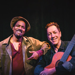 Daniel Carlton and guitarist David Gonzalez smiling and standing side by side against a black stage curtain. Carlton is wearing a tan jacket and a matching hat, and his arm is on Gonzalez's shoulder. Gonzalez is wearing a dark denim jacket and holding his guitar in his arms.