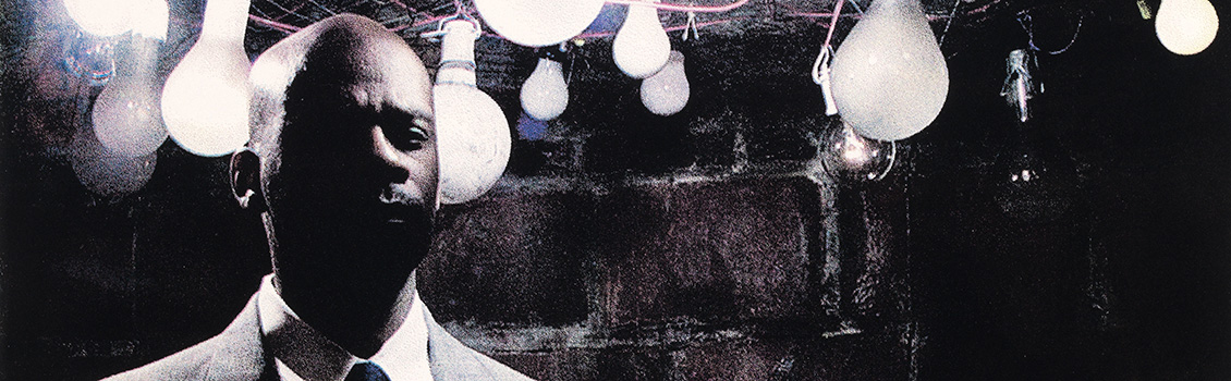 Poto of a man's head in front of light bulbs.  There is a grainy quality and some highlights in pink and blue.  The lower half of the man's face is in shadow.