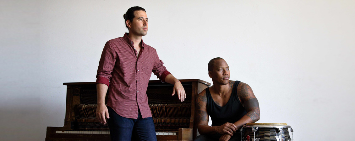 Alfredo Rodriguez and Pedrito Martinez  by a piano and a drum.  Rodrigues is standing, earing a dusty red shirt, he has brown hair. Pedrito is wearing a tank top and bright red hitops. 