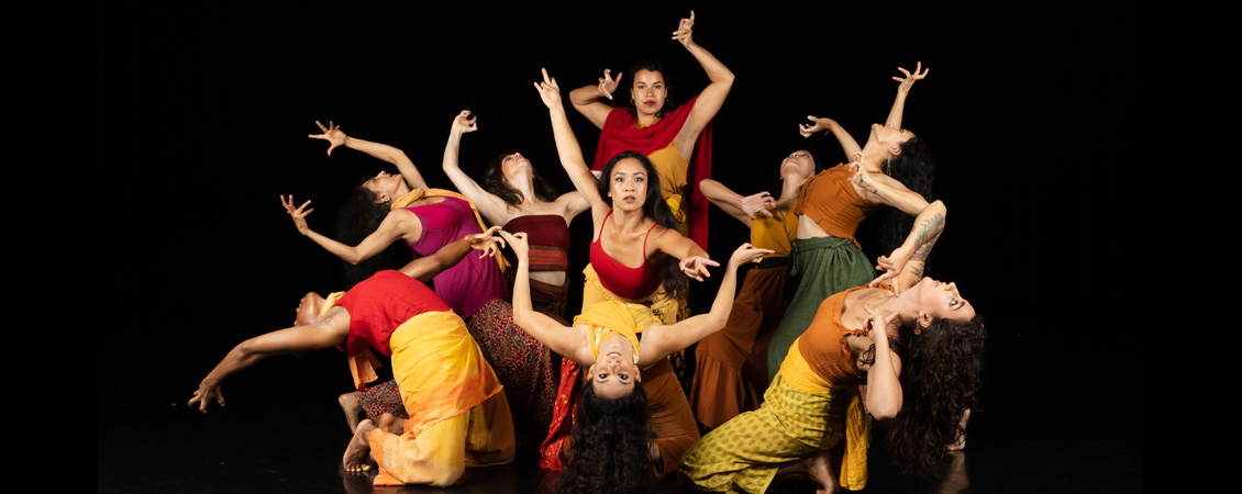 Eight dancers intertwined in an arc on a black background.  They have mostly red, brown and yellow costumes.  The center figure is reaching forward over another in a back bend