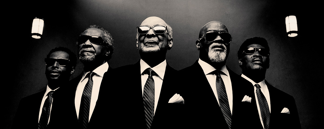 Five black men in suits below a rosette window.  They are all wearing black glasses. The image is in black and white.  