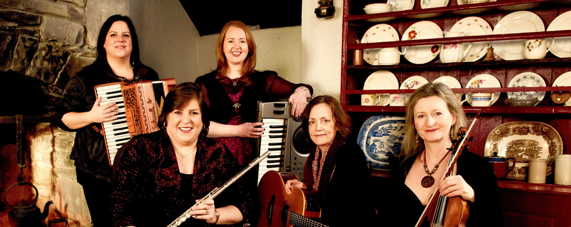 Cherish The Ladies Five women with their instruments in front of a hutch with plates showing.  They women are all in black and holding: accordian, keyboards, flute, guitar and violin.