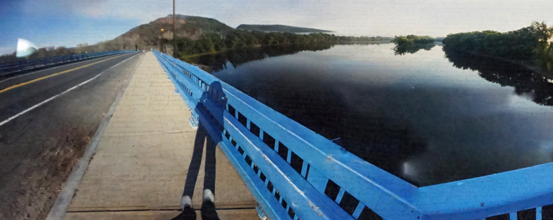 A photo of a brige with blue railings, a river on the right,  and a mountain in the background.   The sky is fairly clear.  The whole photo looks posterized and a little grainy.