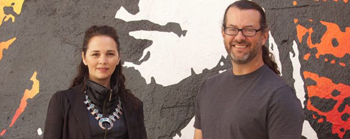  Larrisa FastHorse and Michael John Garcés in front of a painting of a Native American face in grey white and orange yellow.  They are both lookign at the camera and smiling.  