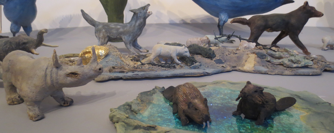 A sculpture depicting a gray wolf howling in front of a cactus with a smaller, while wolf running in front. In front of the wolves there is a giant, blue bird overlooking a brown wolf. A separate sculpture is pictured in front of the first, with it depicting two beavers in a pond. 