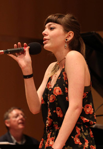 Sepia toned photo of a woman holding a microphone.  She has brown hair with bangs and is wearing a red and black flowered dress