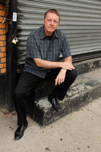 Jeffrey W. Holmes sitting in front of a metal garage door like in a storage unit wear grey and frowning.