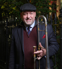 Trombonist Steve Davis holding his instrument wearing a suit, red sweater and cap.  he is in front of some vines