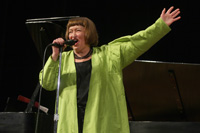 Sheila Jordan with her arm out singing.  She is wearing a lime green duster and is holding the mic in her hand.  She has brown short hair and bangs and is in front of a piano.
