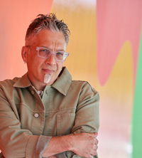 Earl macDonald in front of a pastel colored swoosh background.  He is wearing a tan shirt, clear glasses.  He has gray and black hari with a grey beard which is just a vertical stripe below his chin.