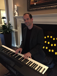 Stephen Page in front of a piano. He is wearing a dark suit and has brown hair and is clean shaven, He is in front of a fireplace with fake candles in it, you can see the corner of a painting's frame on the wall