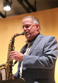 Bruce Diehl playing sax in a grey suit.  He has grey hair and glasses and is in front of a wood wall