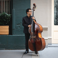 A man in a dark suit standing next to a bass.  He has black hair and glasses. He is in front of a green painted brick and white wall with a planter box.