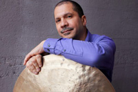 Steve Johns wearing a blue shirt and resting his arm on a huge cymbal.  He has thinning black hair and straggly facial hair