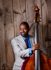 Avery Sharpe in front of a wood wall holding his bass.  He is wearing a blue shirt, red tie and grey suit jacket.  He has medium brown skin and some light facial hair.  He is smiling with mouth closed.