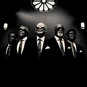 Five black men in suits below a rosette window.  They are all wearing black glasses. The image is in black and white.  
