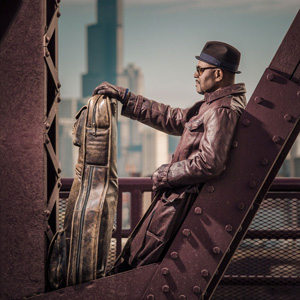 Bobby Broom  leaning on a steel girder holding his instrument case  He is wearing a hat and a dusty burgundy leather jacket. 