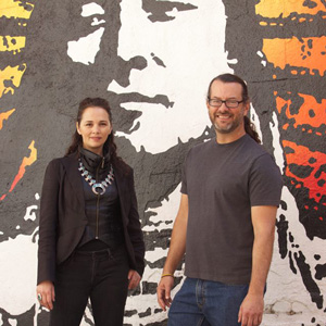  Larrisa FastHorse and Michael John Garcés in front of a painting of a Native American face in grey white and orange yellow.  They are both lookign at the camera and smiling.  