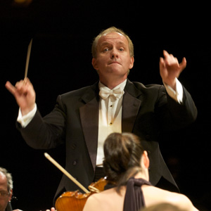 Theodore Kuchar, Conductor holding the baton in his right hand with left in a conductor pose.  He has blond short hair and it wearing a tux with white bow tie