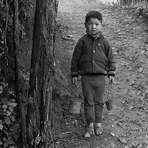 Black and white photo of a boy carrying two buckets.  He is on a path with trees to the left, he is wearing a padded jacket, leggings and sandals