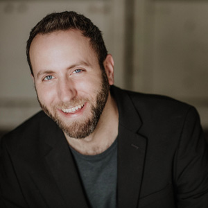 Adam Cicchilitti smiling and facing the camera.  He is wearing a suit, had brown hair, blue eyes and a beard.