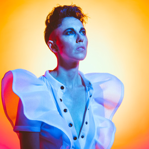 An image of a woman with short hair and dark makeup looking into the distance in blue and pink light. She is wearing a white vest with soft flared shoulders.