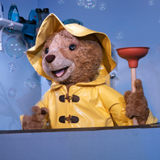 Paddington wearing a yellow rain slicker and holding a red toilet plunger up.