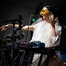 Sirintip, woman wearing yellow sunglasses wearing white shirt seated behind electronic musical equipment on stage. Photo taken at GroundUP Festival by Brian Friedman.